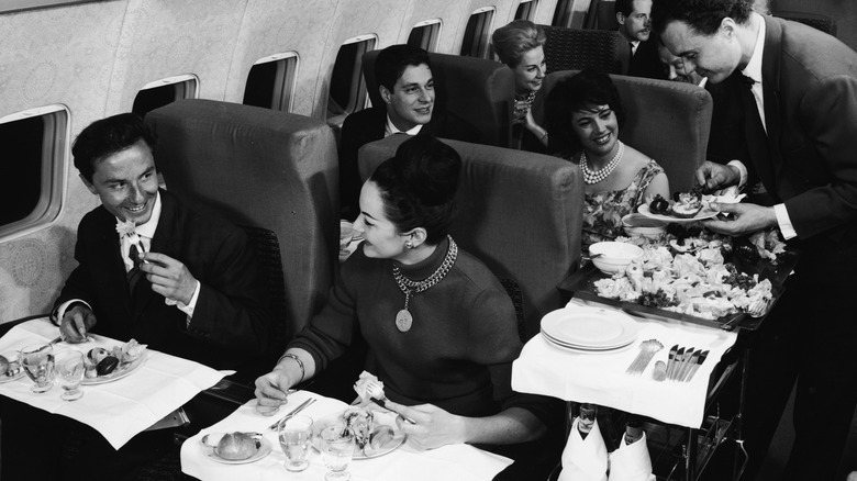 Black-and-white shot of plane passengers being served food by an air steward from a trolley