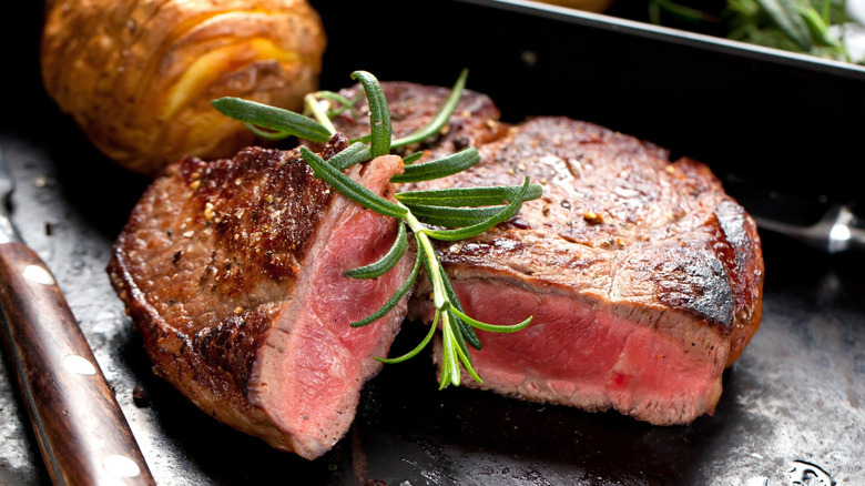 Close up of a steak with a sprig of rosemary on top. A potato and knife handle are in the backdrop.