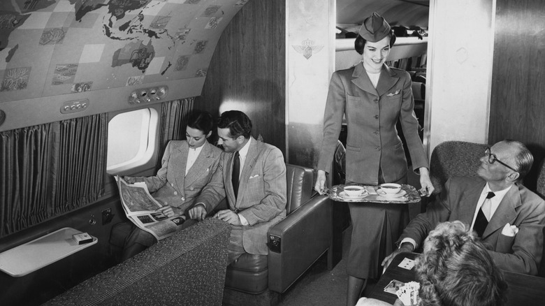 A stewardess in uniform serves tea to a couple in first class, and a couple reads across the aisle.