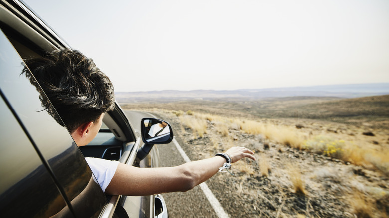 A passenger extends their arm and head out of their car window while cruising down a highway through an open landscape