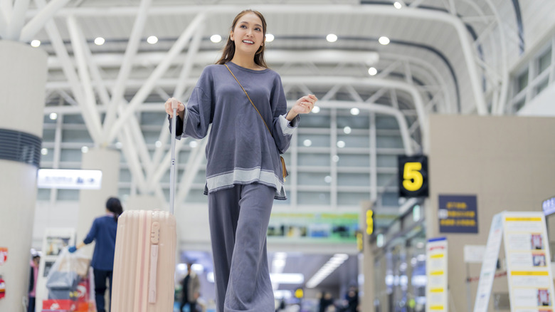 A smiling woman in pajamas strides through an airport with rolling luggage