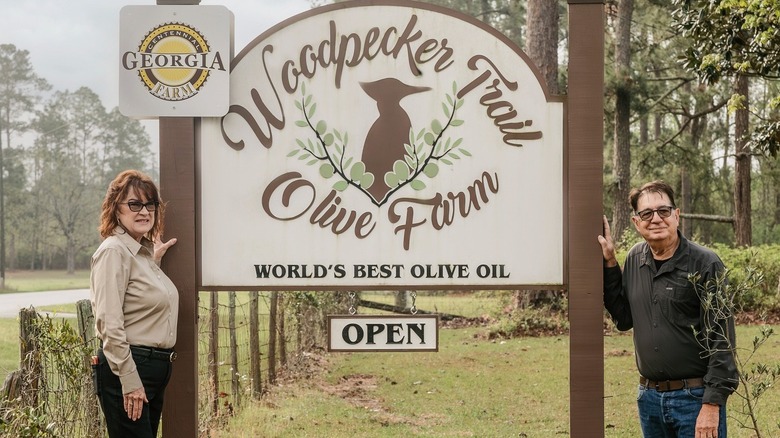 Two people stand next to a sign for an olive farm
