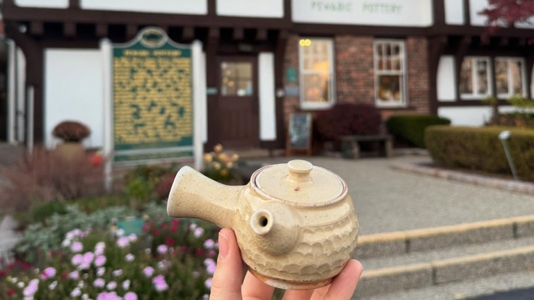 A hand holding a traditional teapot in front of the Pewabic Pottery studio building