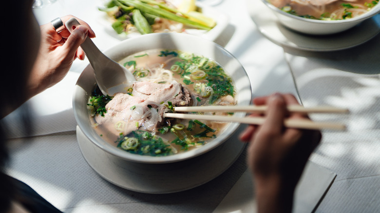 Person eating a bowl of Pho with a spoon and chopsticks