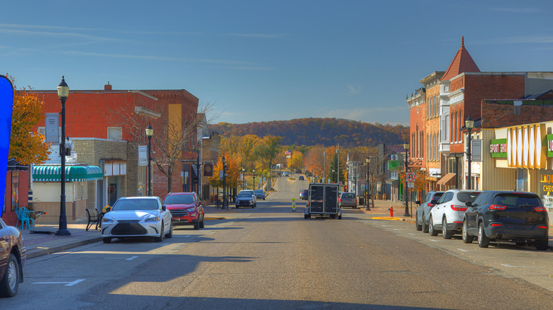 A wide street with parked cars and historical buildings on either side stretches into the distance. The bright blue sky and autumn foliage create a vibrant and inviting atmosphere. Hills can be seen in the background, suggesting a scenic small-town setting. Prairie du Chien, Wisconsin