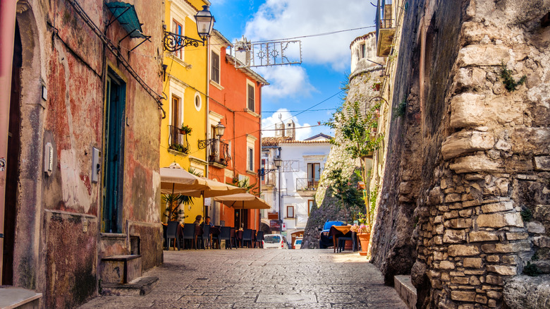 Small alleyway in Vico del Gargano, Italy with old, stone buildings lining the walkway, and three umbrellas outside of a restaurant.