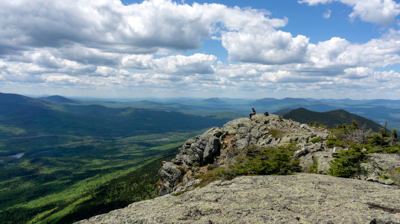 A hiker sits atop a rock outcropping at the summit of Bigelow Mountain Maine with lush forest and mountains cascading in the background.