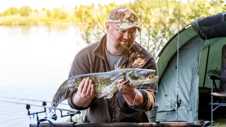 A man in a camo hat holds up a large catfish by the lake