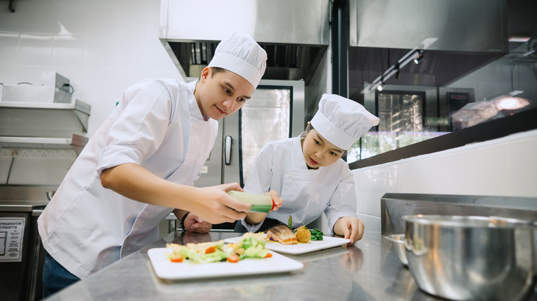 Two culinary students watch carefully while plating a dish of food
