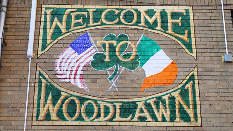A mural on the side of a brick building that reads "Welcome to Woodlawn" with a clover, and American flag and Irish flag all overlapping