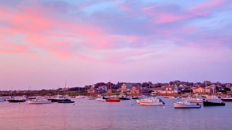 Small boats in the water in front of Little Compton, Rhode Island shoreline with houses lining the water during a colorful sunset.
