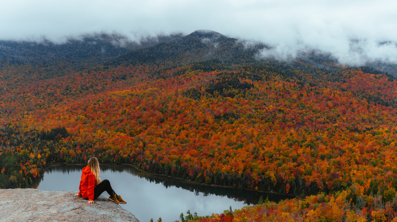 A hiker in a red jacket looks out at the autumn orange trees from the top of Mount Jo