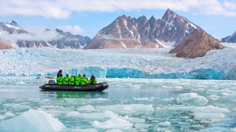 View of a Zodiac boat making its way with its few passengers and the operator through ice floes on a bright day, with a glacier and jagged, snowcapped mountain peaks visible in the background