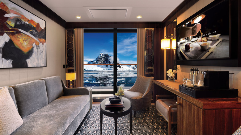 Interior view of the living room, with a plush sofa and chairs, coffee station, TV screen, and veranda, in the Reserve Collection Journey Suite, one of the most luxurious suites aboard the World Navigator, an Atlas Ocean Voyages expedition vessel, with a view of mountain peaks and the ocean outside visible in the background
