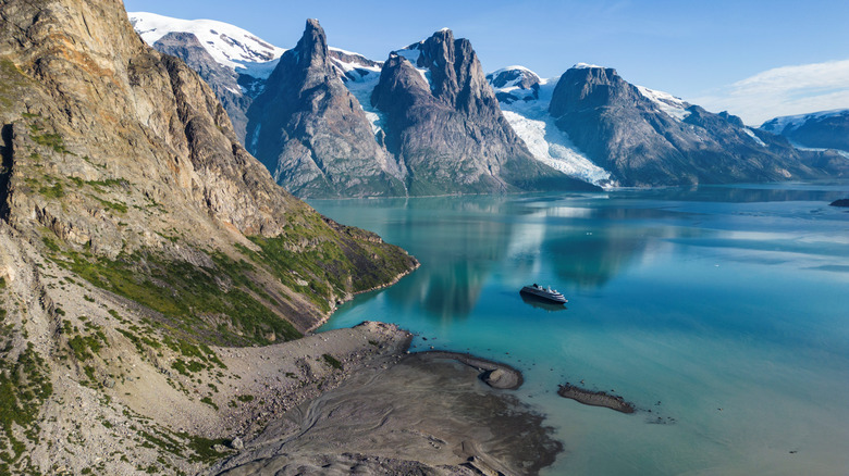 Aerial view of Evighedsfjord in Queqatta, Greenland on a bright day, with jagged, snowcapped peaks in the background and Atlas Ocean Voyages' World Voyager ship visible in the calm waters below