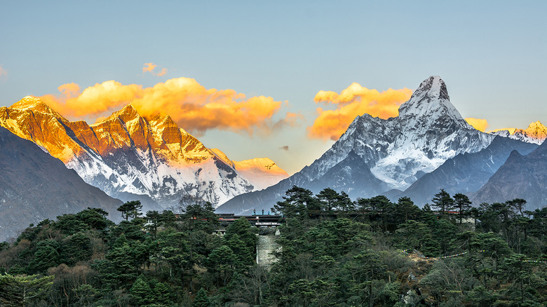 A low-slung building sits atop a forested hill in front of towering ice-covered peaks of the Himalayas