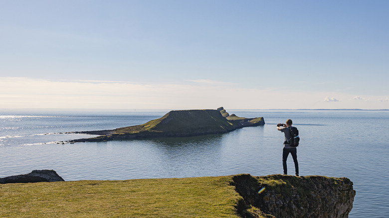 A person looks out at the rugged Worm's Head rock formation just off the cliffs of Llangennith