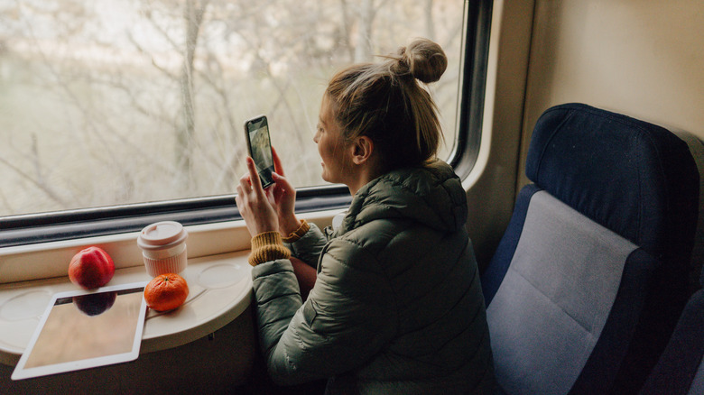 A woman with fruits and coffee next to a train window holding her phone