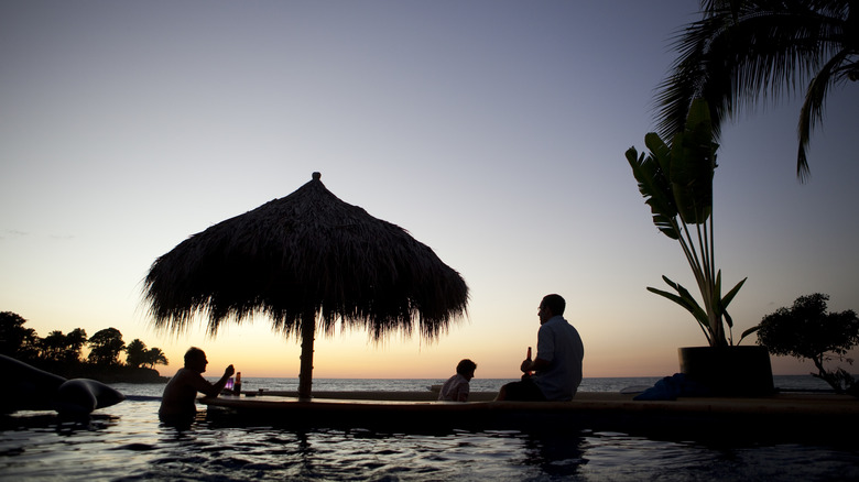 Silhouette of people and a palapa by a pool at sunset