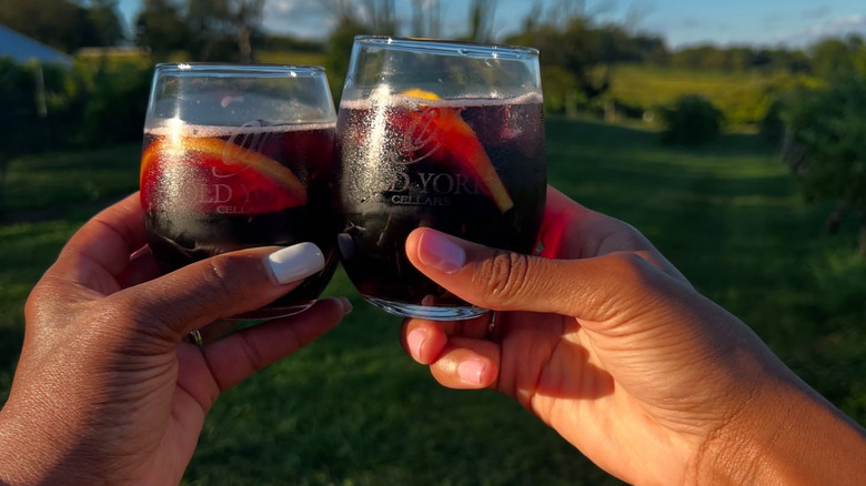 Two hands holding glasses of sangria with green vineyards in the background.