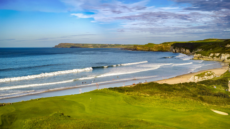 A view of a white sand beach and blue waters past golf courses on a cliff in Portrush, Northern Ireland