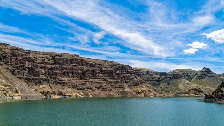 View of a blue lake with rocky desert cliffs in the backdrop under a blue, slightly cloudy sky
