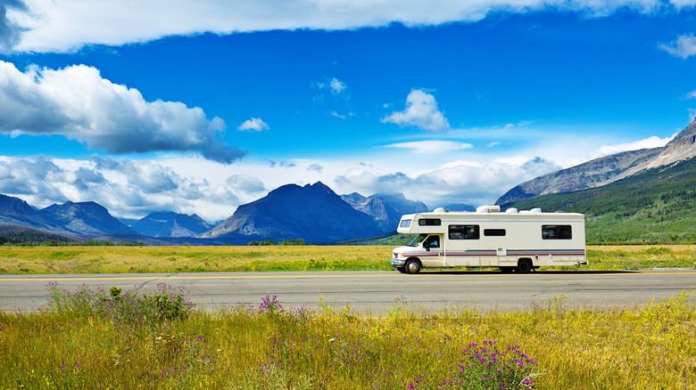 A white RV on an empty road with the backdrop of Glacier National Park mountains and blue cloudy skies
