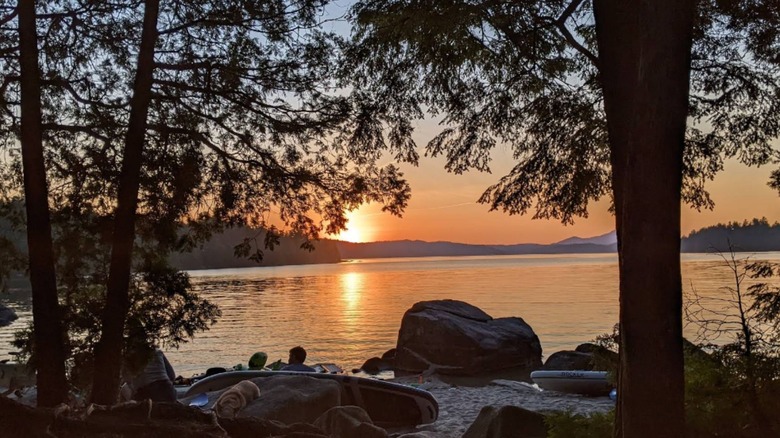 Sunset over a the calm Sebec Lake framed by tall trees, with kayaks and large rocks along the sandy shoreline.