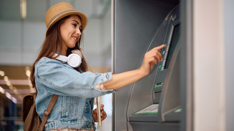 A young, smiling female tourist wearing a strong hat and headphones withdraws money from an ATM at an unidentified airport.