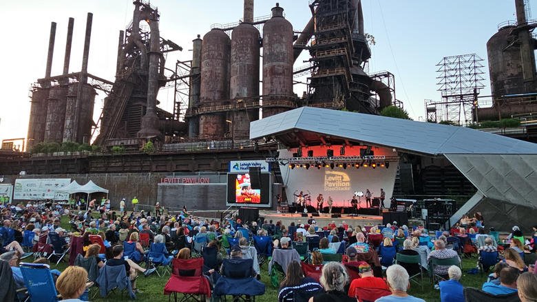 The rusted metal stacks of Bethlehem Steel behind a silver bandshell for Levitt Pavilion, with a rock band on the stage and a crowd of people in folding chairs on the grass in front of it, under a pale blue sky