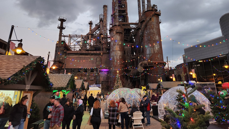 SteelStacks Christkindlemarkt, with a rusting furnace building in the background, wooden vendor booths along the left side, clear domes, crowds, and Christmas trees in the center, and a gray building on the right, under a cloudy sky