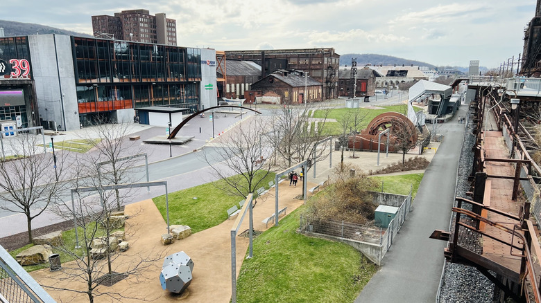 A street in SteelStacks, with a gray building with large windows and abandoned industrial buildings on the right, the furnace walkway on the left, and grass with walkways and statues beside the street, under a cloudy sky