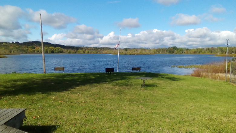 LeBoeuf Lake, a blue lake with green grass in the foreground and three wooden benches along the shore and trees along the far shore, under a cloudy blue sky