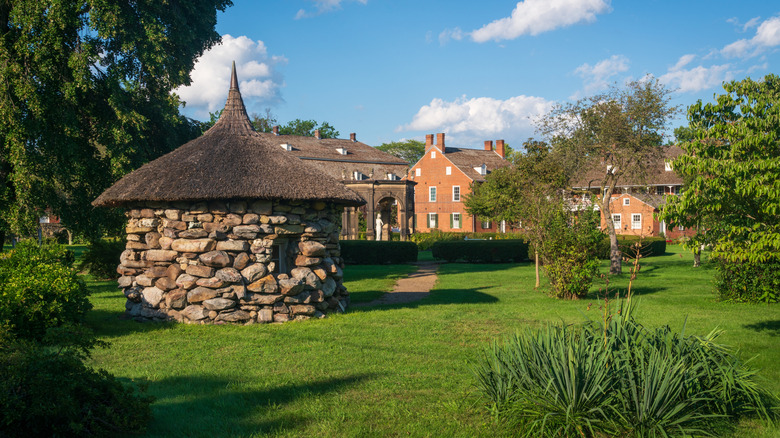 Shot of Old Economy Village, with a round stone structure in the foreground surrounded by grass and trees, with brick buildings in the background under a blue sky