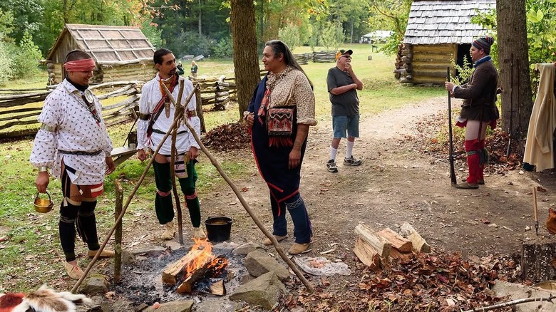 Reenactors dressed in Native American attire stand around a fire at Meadowcroft Historic Village