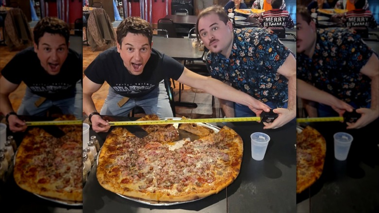 Two men with a very large pizza and a tape measure