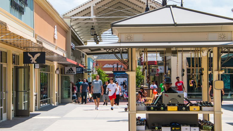 Gloucester Premium Outlets, with shoppers walking by a peach-walled building with stores and a kiosk in the foreground