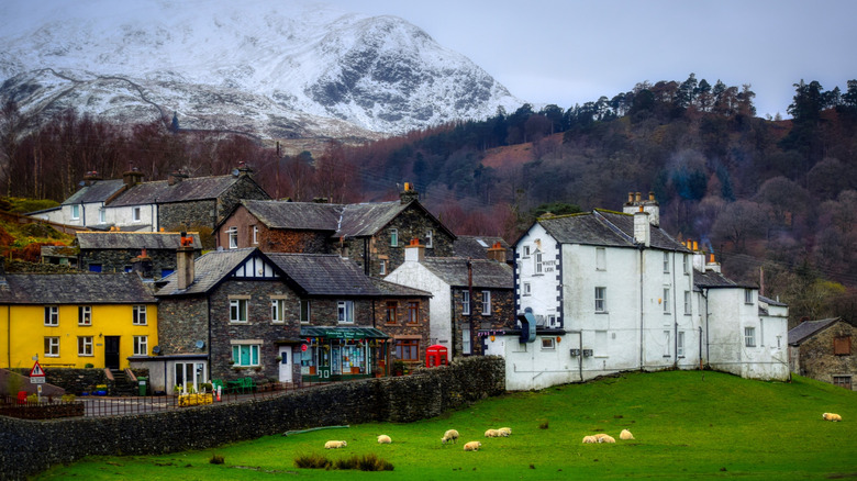 Brick and colored buildings behind a meadow and stone wall in Patterdale, with forests and snow capped mountains behind it