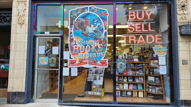 Front of Amazing Books and Records, with large windows displaying a colorful store sign and red-lighted words reading "BUY SELL TRADE" and shelves of books visible inside the store