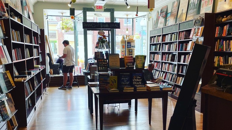 Inside Penguin Bookshop, with shelves of books along each wall and a table of books in the center of a wooden floor, one shopper crouched beside the shelf on the right, and another in a white shirt standing by the front window