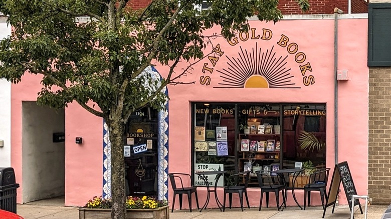 Front of Stay Gold Books, a pink building with white and blue tiles around the door and black tables and chairs out front of a large window displaying books, with a tree and flower box in the foreground