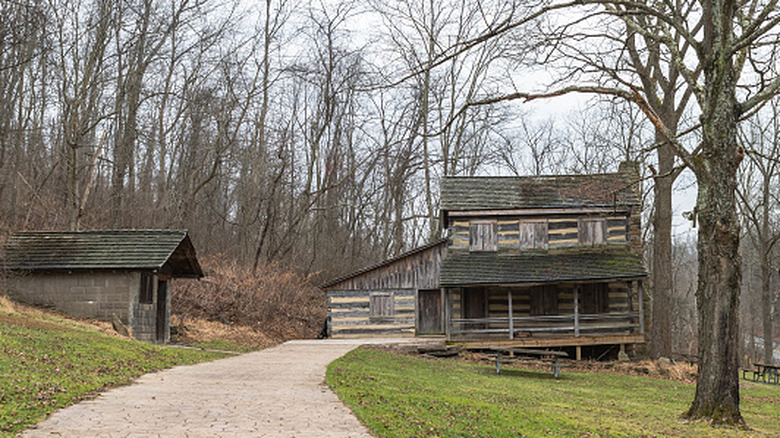 Carpenter Log Cabin, a two-story wooden building beside a paved path on the right, with a smaller stone structure on the left, surrounded by green grass, winter trees, and picnic tables