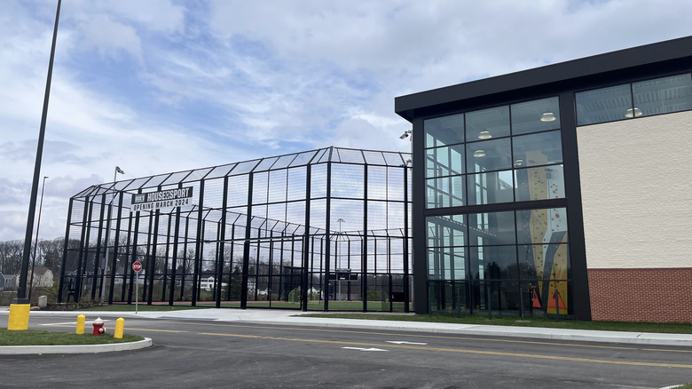 Black fence around a sports field, next to a two-story black-framed window with a climbing wall inside, with a parking lot in front under a cloudy blue sky
