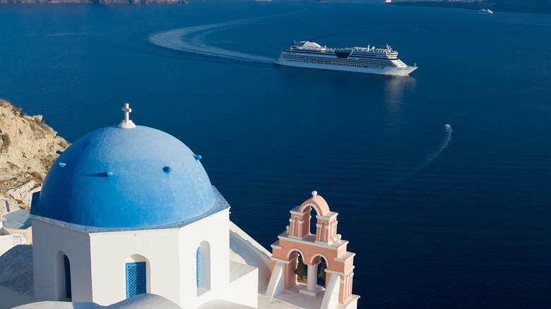 A cruise ship gliding through the water in front of a blue church dome and peach-colored bell tower in Santorini, Greece