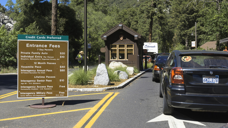 Cars wait in line to pay entry at a Yosemite toll booth beside a sign detailing ticket prices