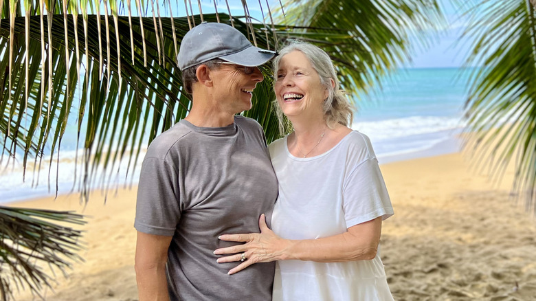 An elderly couple smiling on a beach under palm trees