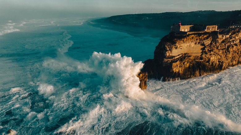 Big ocean waves crash against a rugged cliff topped with a historic red and white lighthouse in Nazaré