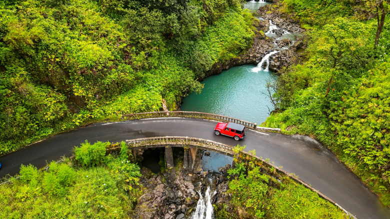 A red jeep driving over a bridge on a Hawaiian road in Maui, surrounded by lush vegetation