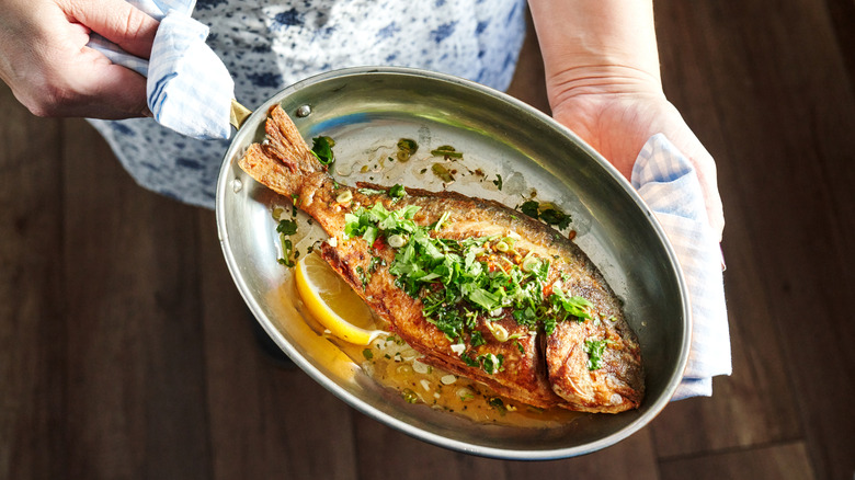 A woman holding a pan of grilled seabeam with lemon slices and garnish