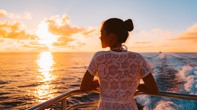 A woman leans against the railing on the deck of a cruise ship at sunset